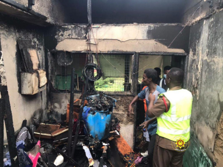Post-fire scene inside a burnt room with charred walls and debris; responding personnel in a high-visibility vest assess the damage.
