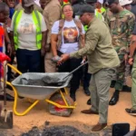 Group of workers and officials surround a wheelbarrow of asphalt; a man in a green shirt shovels material during a road repair scene, with several people wearing high‑visibility vests.