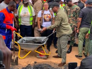 Group of people around a wheelbarrow with wet cement as a man shovels at a ground‑breaking or construction event.