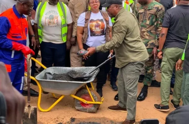 Group of workers and officials surround a wheelbarrow of asphalt; a man in a green shirt shovels material during a road repair scene, with several people wearing high‑visibility vests.