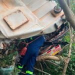 Emergency responder in blue with high‑visibility stripes searches under an overturned truck tangled in branches.
