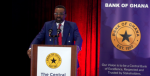 A man with glasses and a beard in a navy suit delivering a speech at a wooden podium with the Bank of Ghana seal, supported by red curtains in the background.