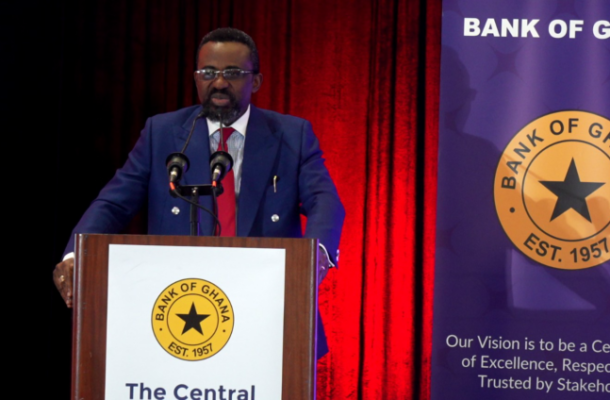 image-298 | Ghanaguardian.com A man with glasses and a beard in a navy suit delivering a speech at a wooden podium with the Bank of Ghana seal, supported by red curtains in the background.