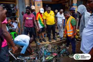A group of people standing around a large pile of garbage on a street, inspecting waste as part of a community cleanup effort, with Accra Metropolitan Assembly logo visible in the corner.