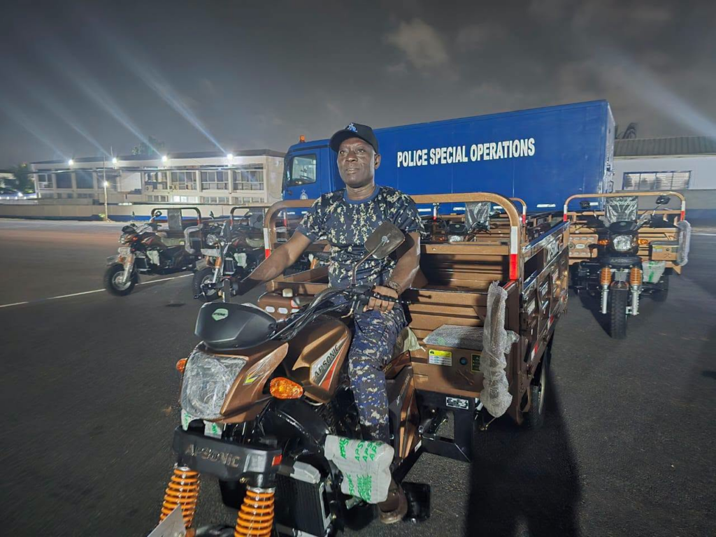 Police officer sits on a brown utility motorcycle with a cargo bed, at night, in a lot near a blue Police Special Operations truck.