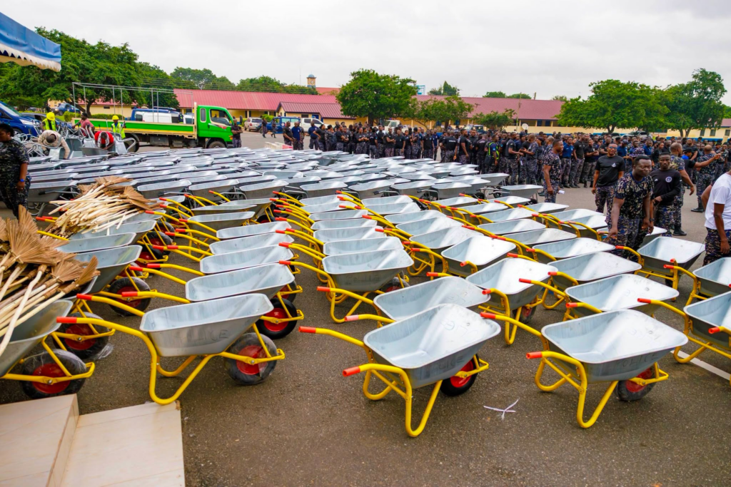 Rows of new metal wheelbarrows with yellow frames lined up on a paved surface, with uniformed personnel in the background at a ceremony or event.