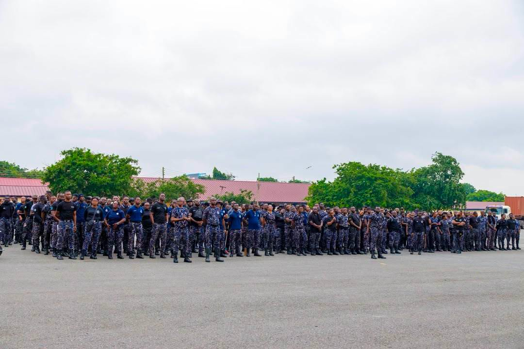 A large group of uniformed personnel standing in formation on a paved lot with trees and red-roofed buildings in the background.