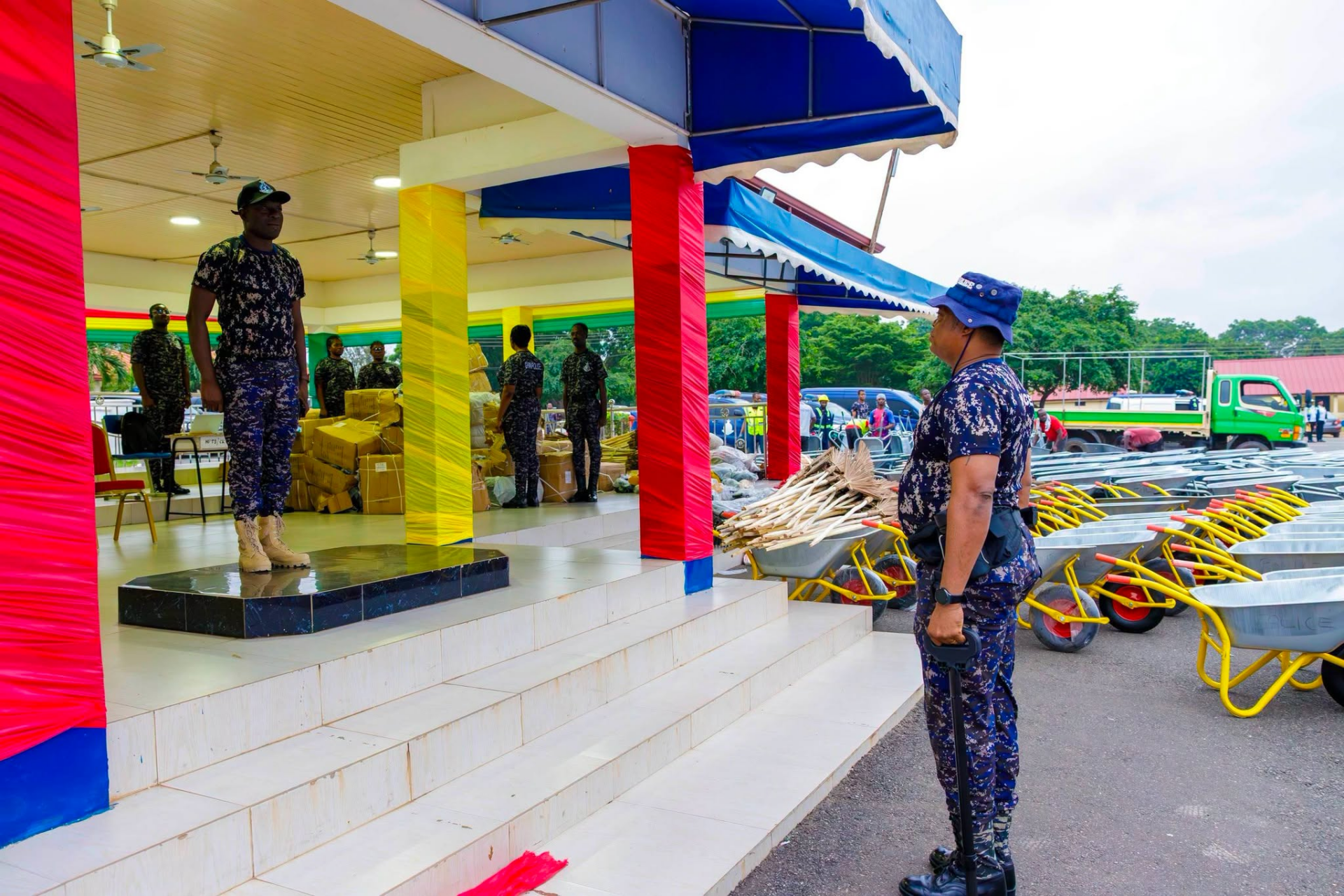 Soldiers in camouflage on a decorated stage with stacked boxes and relief supplies at an outdoor aid event.