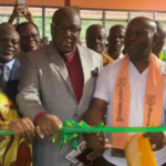 People attend a ribbon-cutting ceremony as a man in a gray suit cuts a green ribbon with another man in a white shirt and orange sash, while onlookers cheer.