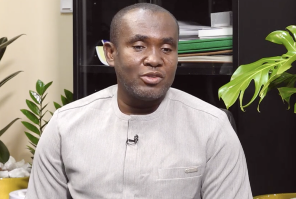 Man seated indoors in a light gray traditional shirt with a lavalier mic, speaking during an interview, plants and shelves in the background.