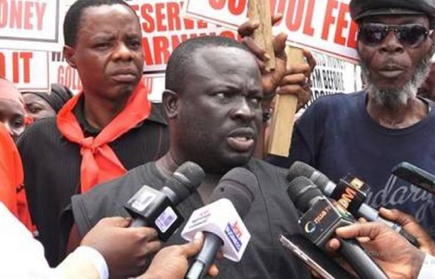 Male speaker in black shirt addressing reporters with multiple microphones, protesters and sign boards visible in the background.