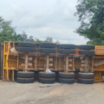 Yellow truck trailer overturned on its side in a yard, with wheels stacked along the frame.