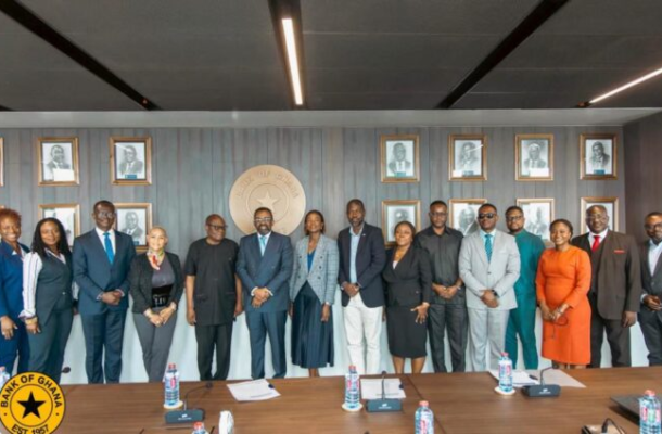Group of 15 professionals in business attire posing for a photo in a conference room, Bank of Ghana seal on the wall with framed portraits behind, table with documents and water bottles in front.