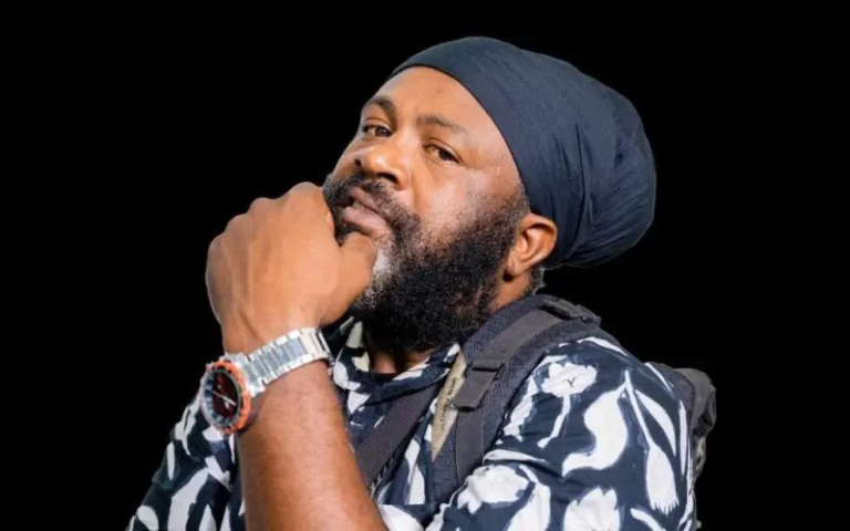 Bearded man wearing a dark blue head wrap, resting his chin on his hand, in a black studio background with a patterned shirt and watch visible.