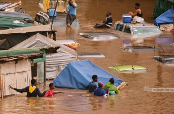 Flooded street with submerged cars and people wading through brown water; tents and shelters line the area amidst the flood.
