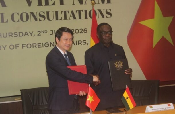 Two officials in suits shake hands after signing documents at a formal diplomatic ceremony, with Vietnam and Ghana flags on the table behind them.