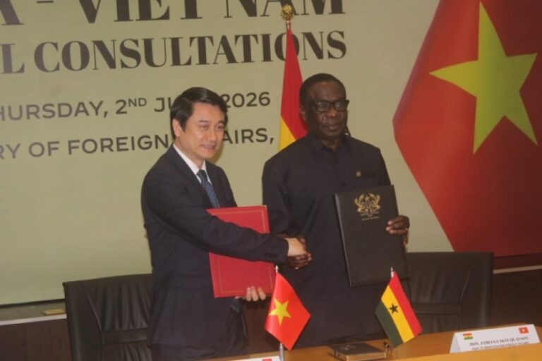 Two officials in suits shake hands after signing documents at a formal diplomatic ceremony, with Vietnam and Ghana flags on the table behind them.