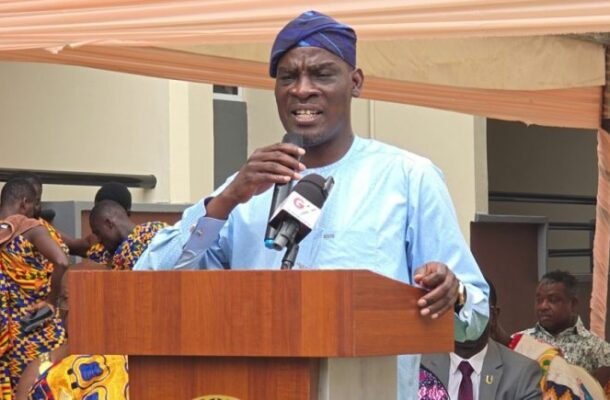Man in a blue traditional outfit and headwrap speaks into a microphone from a wooden podium under a tan canopy at an outdoor event.
