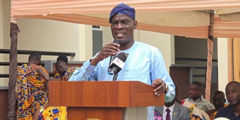 Man in a blue traditional outfit and headwrap speaks into a microphone from a wooden podium under a tan canopy at an outdoor event.