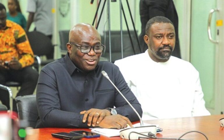 Two men sit at a conference table; the man in black speaks into a microphone while the other listens.
