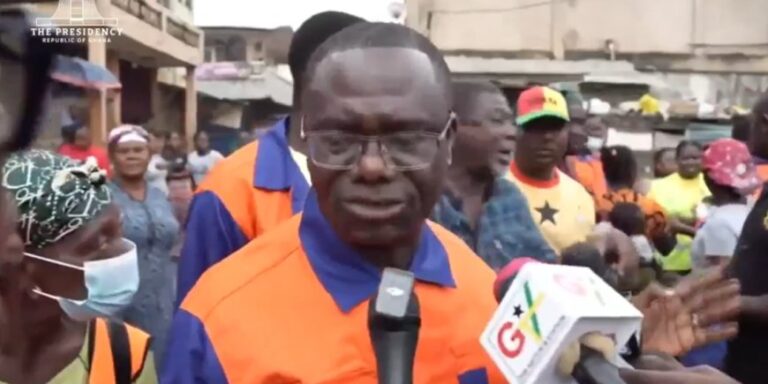 Man in an orange safety vest being interviewed by reporters in a crowded outdoor area; a microphone with a G-mark is held close to him.