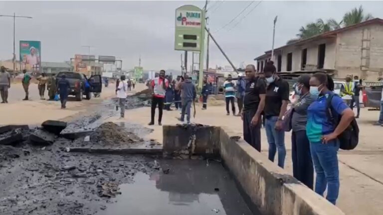 A group of bystanders and responders stand along a collapsed road with a large sinkhole and wet, charred debris, as a car and construction activity are visible in the background.