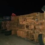 Overturned large truck on its side at night, with emergency responders and a police vehicle with green lights nearby.