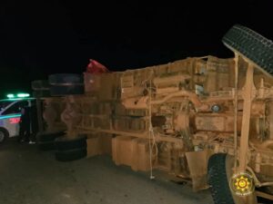 Overturned large truck on its side at night, with emergency responders and a police vehicle with green lights nearby.