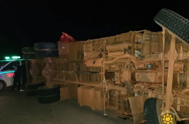 Overturned large truck on its side at night, with emergency responders and a police vehicle with green lights nearby.