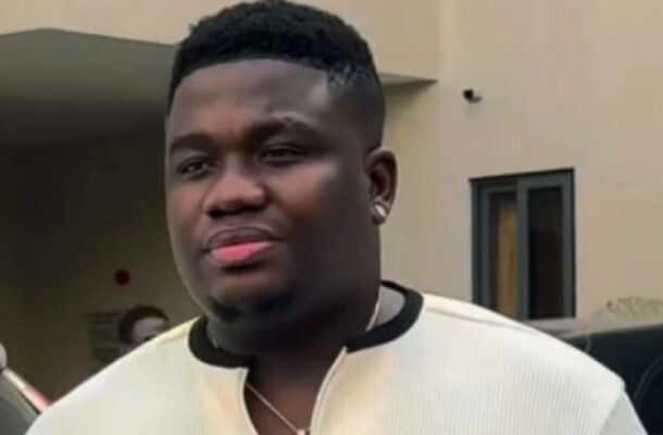 Man outdoors wearing a light beige/cream long-sleeve shirt with a black collar trim, looking slightly to the left, in front of a building and parked cars.