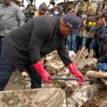 Man in dark clothing and red gloves pries apart rubble at a debris pile while a crowd of reporters films nearby.