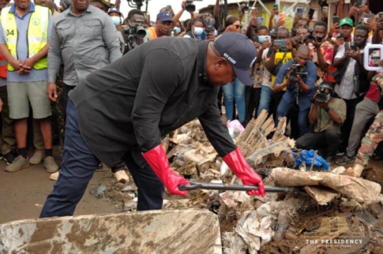 Man in dark clothing and red gloves pries apart rubble at a debris pile while a crowd of reporters films nearby.