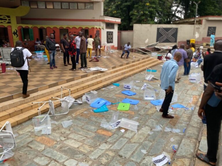 Outdoor scene with a group of people standing on a stepped terrace as debris and scattered plastic chairs cover the surrounding walkway, suggesting a disrupted or abandoned event.