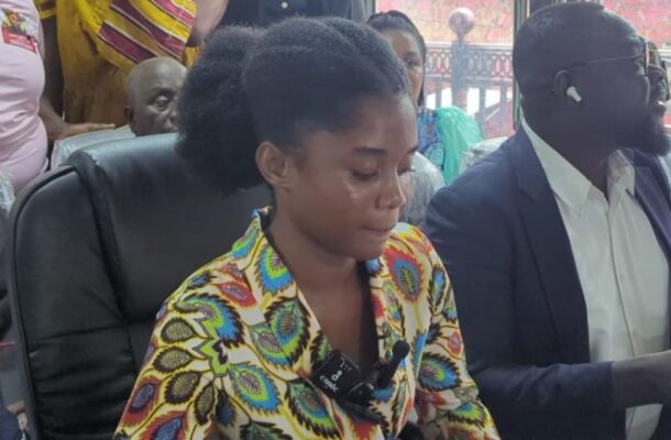 Woman in a vibrant, patterned blouse sits at a conference table with a lapel microphone clipped to her chest, others seated behind her in a meeting setting.