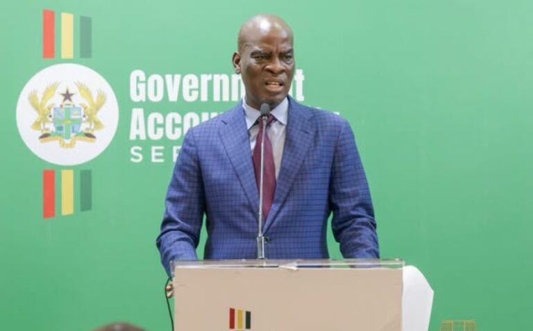 A man in a blue suit speaks at a podium on a green backdrop with a government emblem behind him.