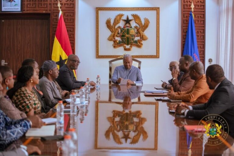 A formal meeting around a long glass conference table with a central official signing or reviewing documents, flanked by colleagues on both sides, in a room with a large framed national emblem and flags in the background.