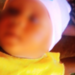 Blurry close-up of a baby wearing a white hat and yellow bib, lying on a brown surface nearby.