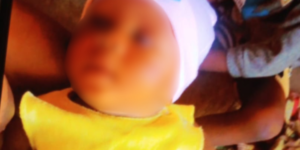 Blurry close-up of a baby wearing a white hat and yellow bib, lying on a brown surface nearby.