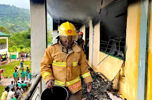 leklebi shs fire 3 | Ghanaguardian.com Firefighter in yellow turnout gear walks along a smoke-darkened balcony, holding a black bucket amid charred debris after a building fire.