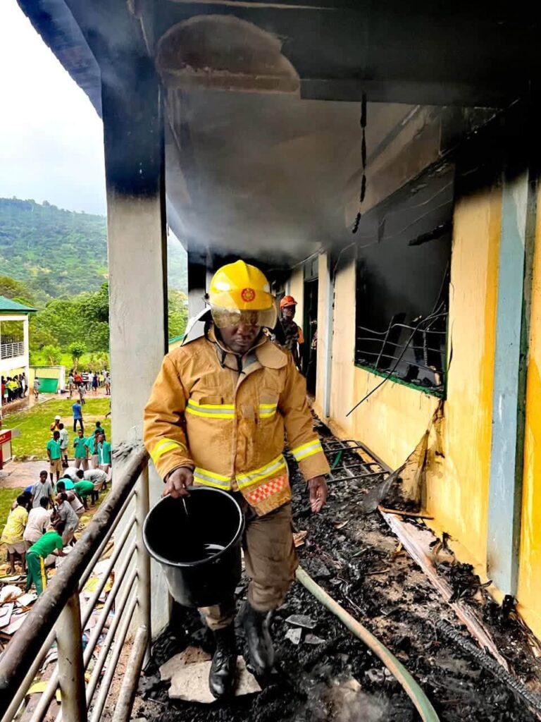 Firefighter in yellow turnout gear walks along a smoke-darkened balcony, holding a black bucket amid charred debris after a building fire.