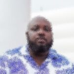 Bearded man in a light blue patterned shirt seated indoors, facing the camera.