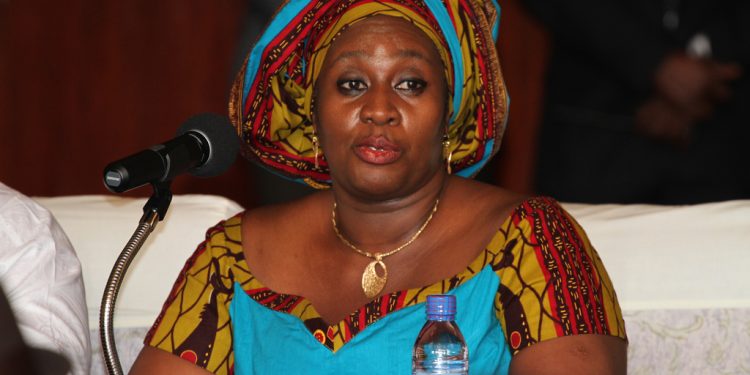 Woman wearing a colorful headwrap and patterned dress sits at a table with a microphone in front, speaking at an event.