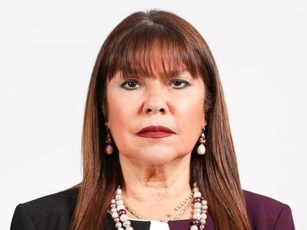 Head-and-shoulders portrait of a serious-looking middle-aged woman with auburn hair and bangs, wearing a dark blazer and pearl jewelry, facing the camera