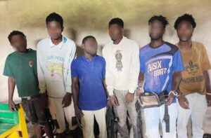 Seven young men standing shoulder to shoulder indoors against a yellow wall; faces blurred, wearing casual t‑shirts and shorts, some with bags.