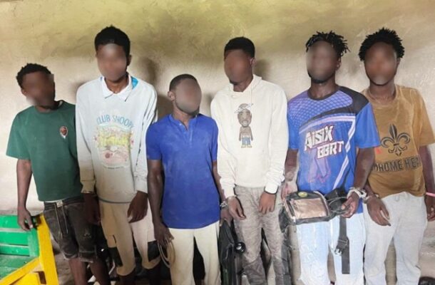 suspects2 | Ghanaguardian.com Seven young men standing shoulder to shoulder indoors against a yellow wall; faces blurred, wearing casual t‑shirts and shorts, some with bags.