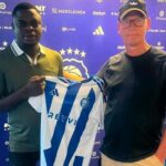 Two men pose with a blue-and-white striped soccer jersey in front of a purple backdrop covered with sponsor logos.
