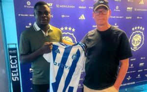 Two men pose with a blue-and-white striped soccer jersey in front of a purple backdrop covered with sponsor logos.
