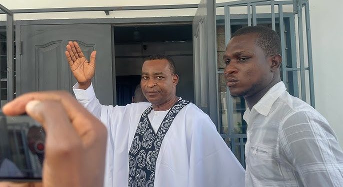 Man in white robe with a patterned stole waves from a doorway as another man in a light shirt stands nearby.