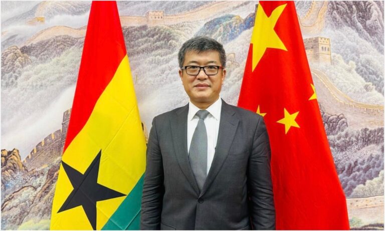 Formal portrait of a man in a dark suit and tie standing between the Ghana flag (left) and the China flag (right).