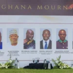 Row of head-and-shoulders portraits on a Ghana Mourns memorial banner at an outdoor ceremony with flowers in front.